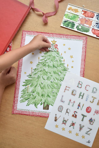 Child's hand placing "A" sticker at the top of The Letter Nest's Advent Alphabet Calendar