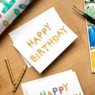 Flatlay styled photo of a Happy Birthday Cards featuring gold balloon letters painted by Sally of The Letter Nest, styled with birthday candles, watercolor paints, and other birthday items from The Letter Nest