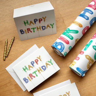 Flatlay styled photo of The Letter Nest's Happy Birthday Cards, shown flat, folded, and alongside the matching Happy Birthday Gift Wrap, all featuring hand-painted watercolor balloon letters in bright color's for a child's birthday