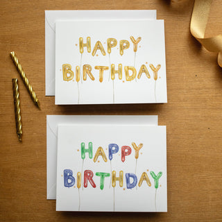 Flatlay styled photo of The Letter Nest's Happy Birthday Cards, shown in gold and multicolor colorways, featuring hand-painted watercolor balloon letters in bright color's for a child's birthday