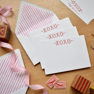 Flatlay styled photo of The Letter Nest's hand-painted, printed watercolor cards featuring stylized "XOXO" Letters at the top, with pink stripe envelopes